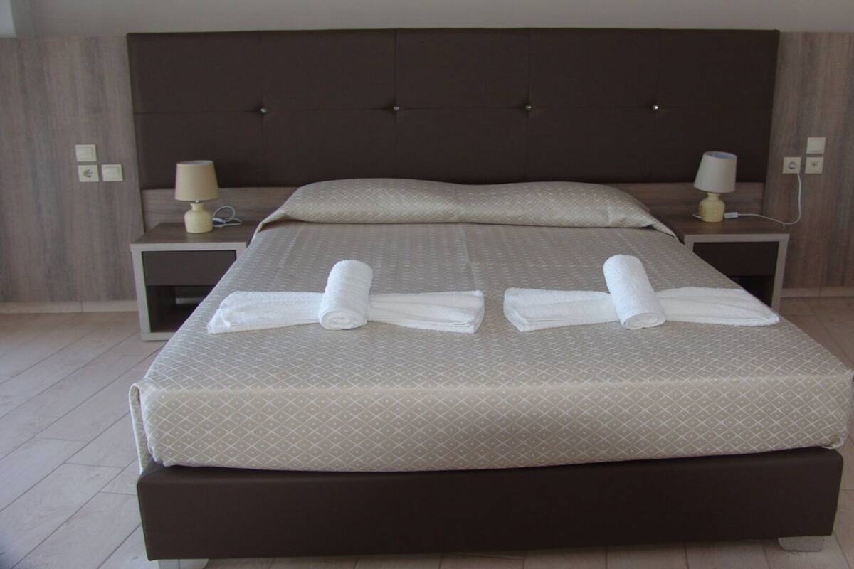 Cama bem arrumada com colcha estampada e duas toalhas enroladas. Há duas mesinhas de cabeceira com abajures e um ponto de luz fixado na parede.