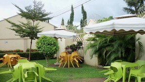 Un espace extérieur aménagé en terrasse, avec des chaises en plastique vertes et jaunes, un petit arbre et un coin salon abrité.