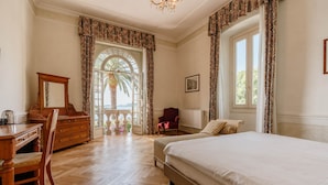 Un dormitorio con una cama grande, un aparador de madera, una silla y un escritorio. Hay una ventana con cortinas y vistas a palmeras.