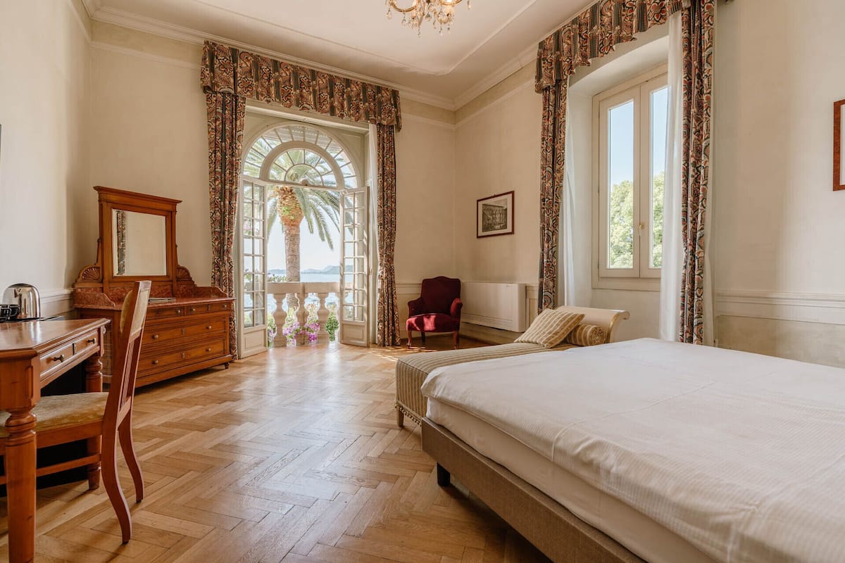 A bedroom with a large bed, a wooden dresser, a chair, and a desk. There is a window with curtains and a view of palm trees.
