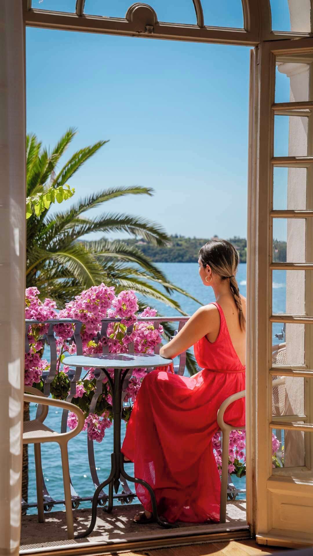Una mujer con vestido rojo sentada a una mesa con flores rosas, con vista a un cuerpo de agua y palmeras.