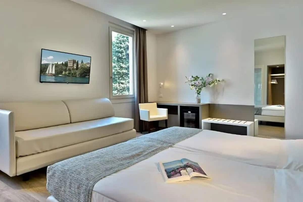A modern hotel room with a bed, a sofa, a desk, and a large window.