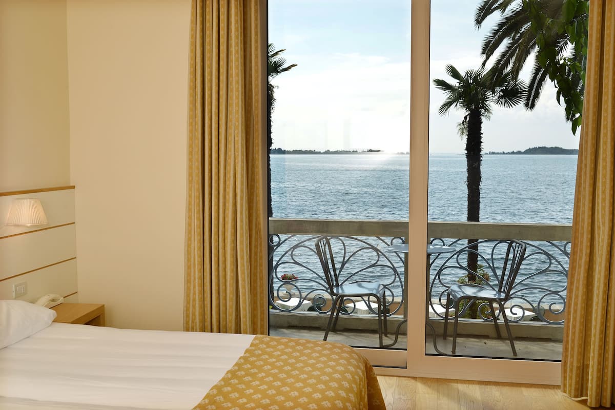 A hotel room with a bed, a bedside table with a lamp, and a view of the sea through a balcony with a metal railing.