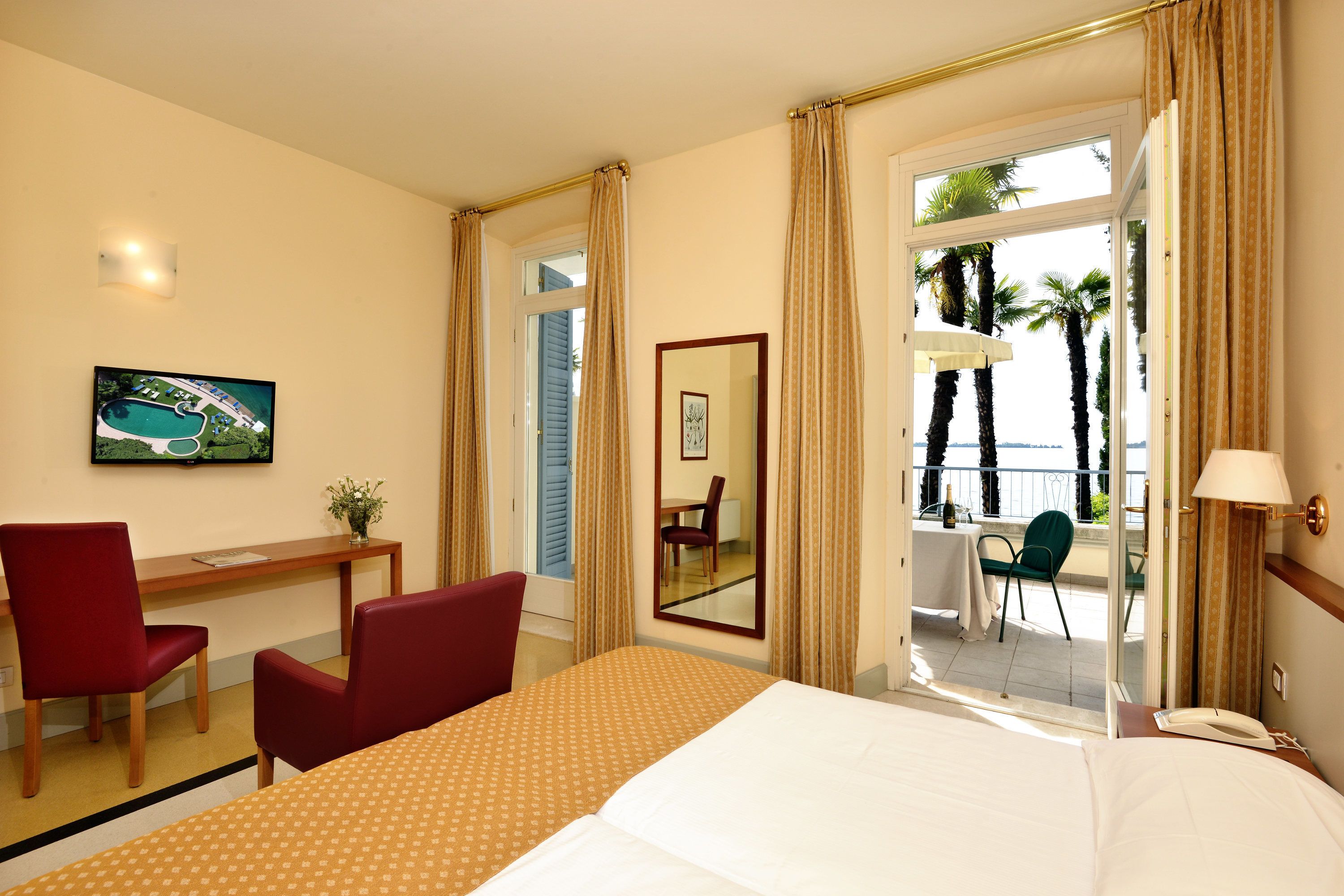 Quarto de hotel com uma cama, uma escrivaninha, duas cadeiras, um espelho e uma varanda com vista para palmeiras.