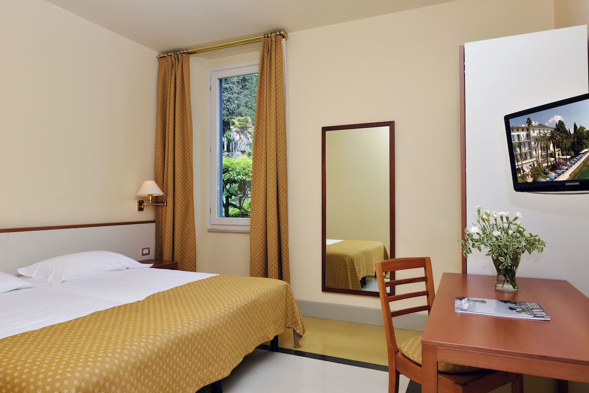 A hotel room with a bed, a desk, a chair, a mirror, a window with curtains, and a wall-mounted TV.