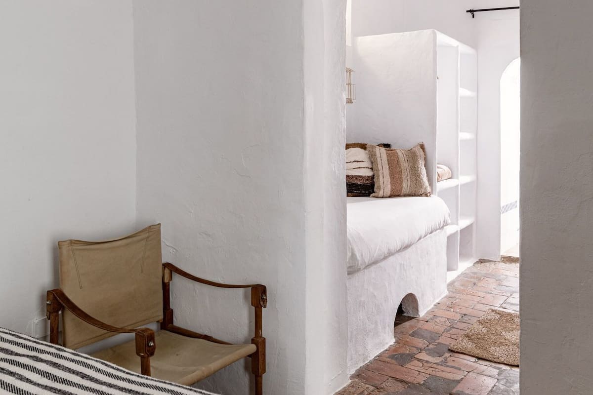 A bedroom with a bed, a wooden chair, and a view through an archway to another room with a bed and a window.