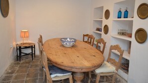 Una mesa de comedor de madera con un tazón, rodeada de sillas, en una habitación con paredes blancas y estantes con platos decorativos.
