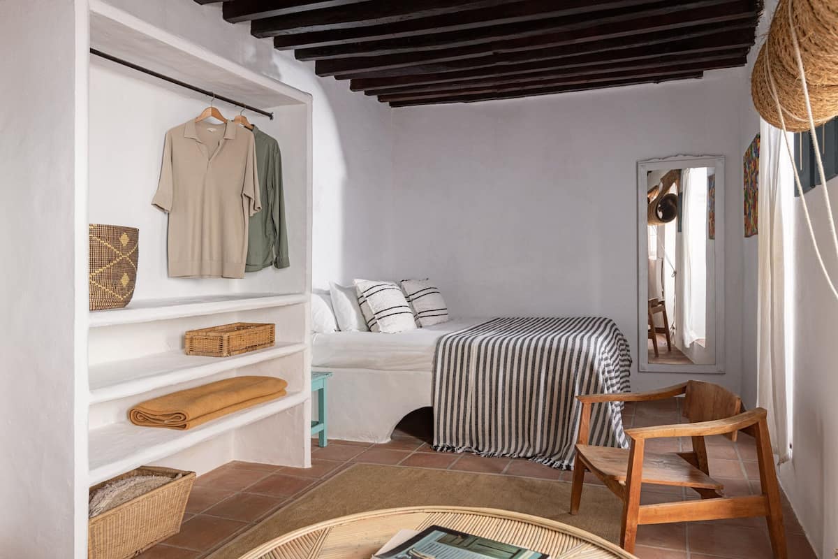 A bedroom with a bed, a wooden chair, a shelf with clothes and baskets, and a mirror.