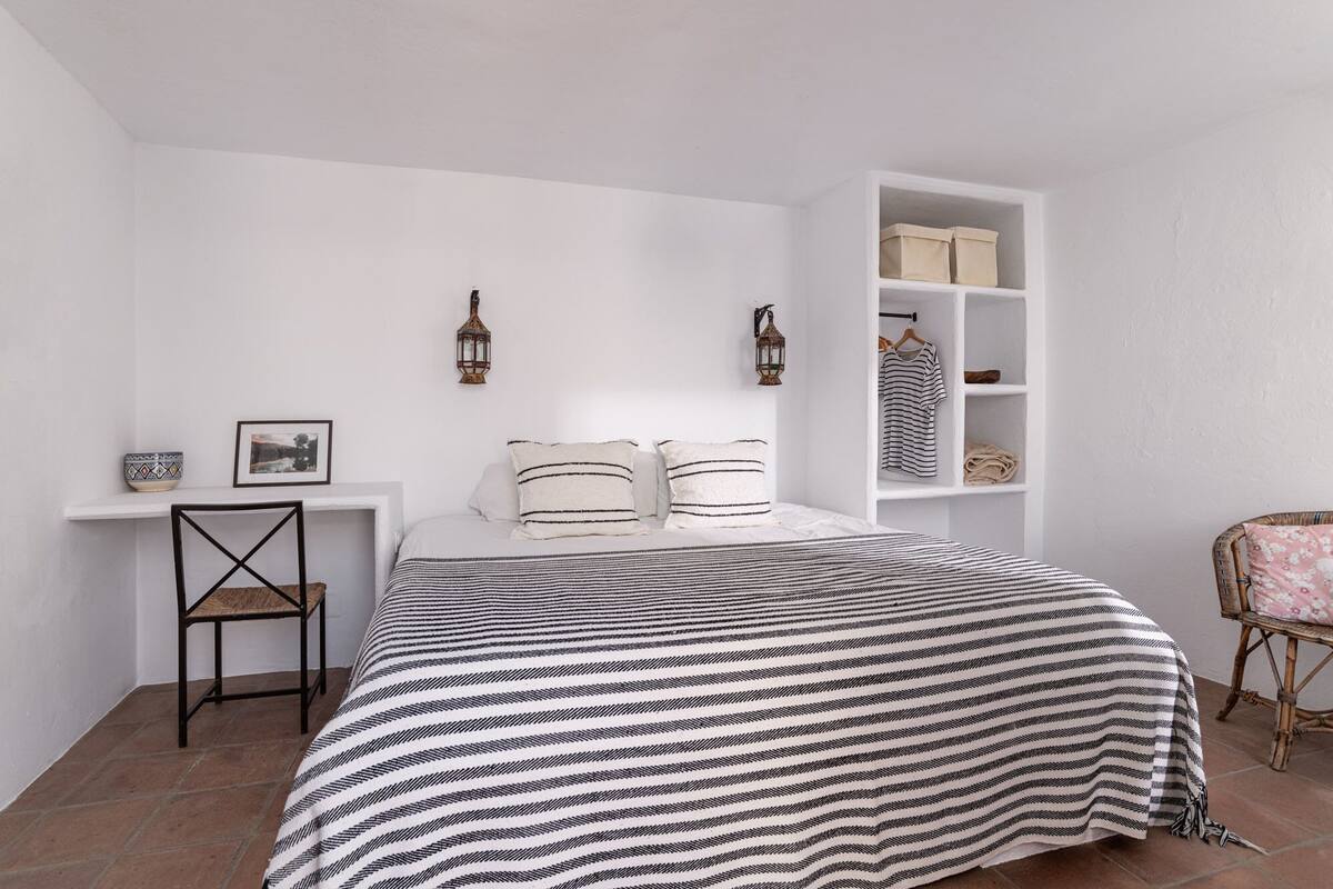 A bedroom with a bed, a chair, a shelf with storage boxes, and two wall-mounted lanterns.