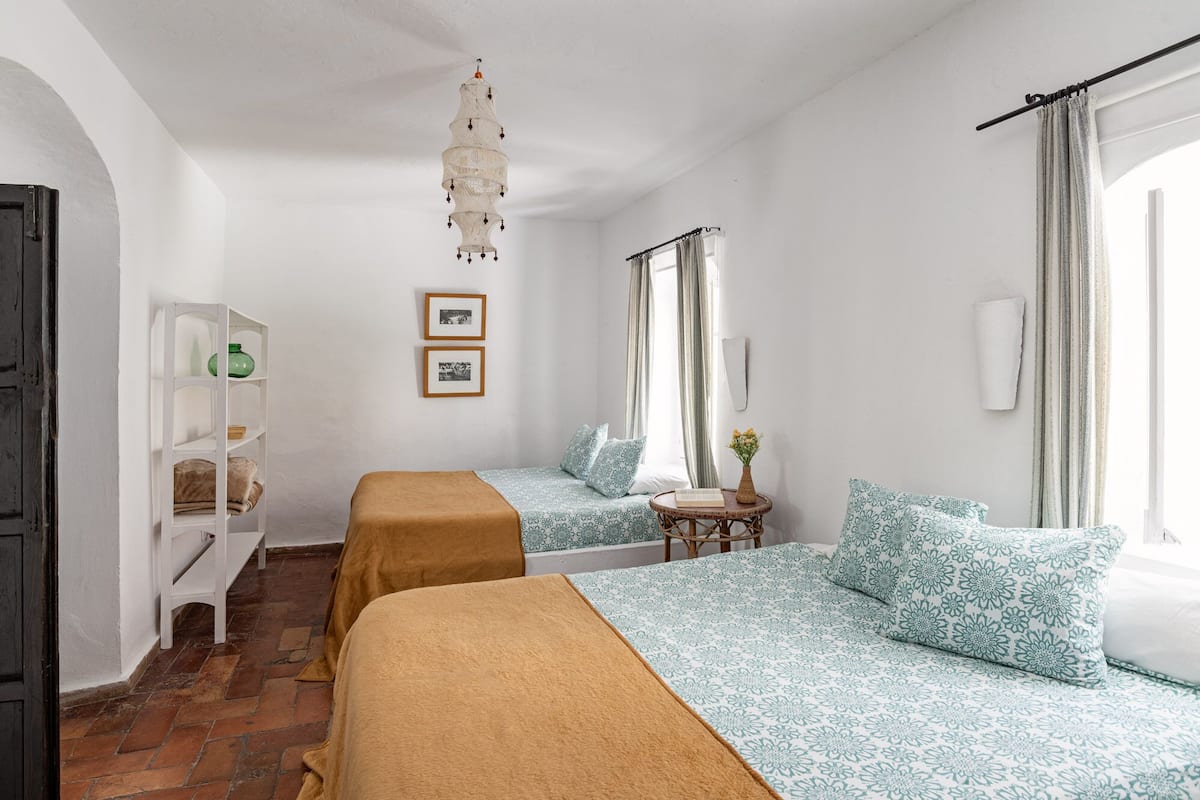 A room with two beds, a hanging light fixture, a small table, and a shelf with items.
