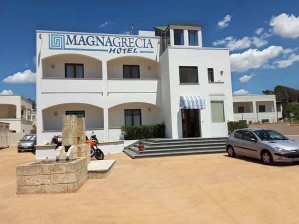 Hotel MAGNAGRECIA de dues plantes amb entrada central, una petita zona d'estar a l'aire lliure i un aparcament.
