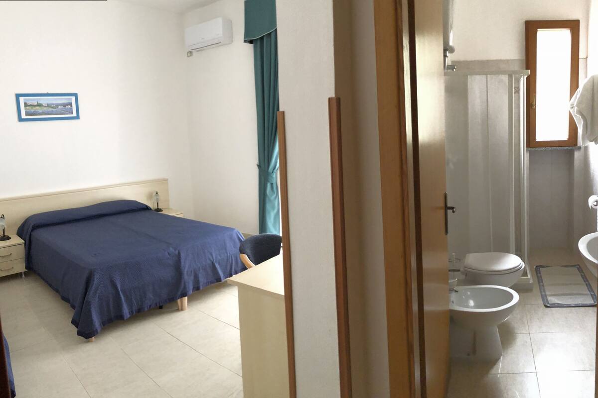 A room with a bed, a bedside table, a desk, a chair, a bathroom with a toilet, a sink, and a shower.