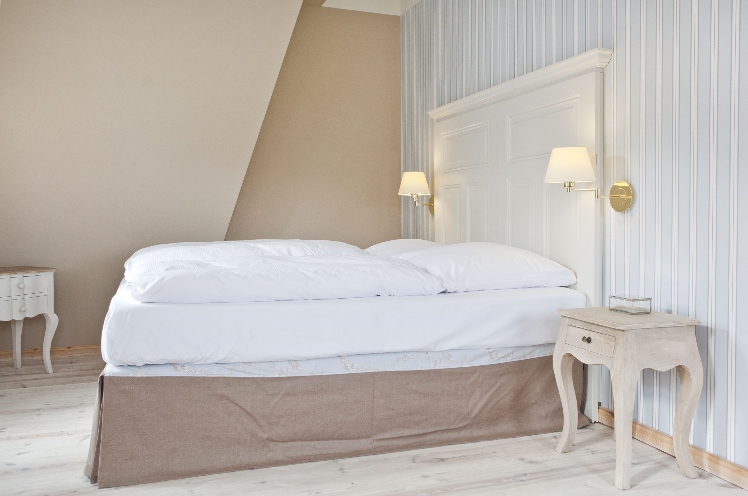 A neatly made bed with white linens, a small bedside table with a lamp, and a wall-mounted mirror.