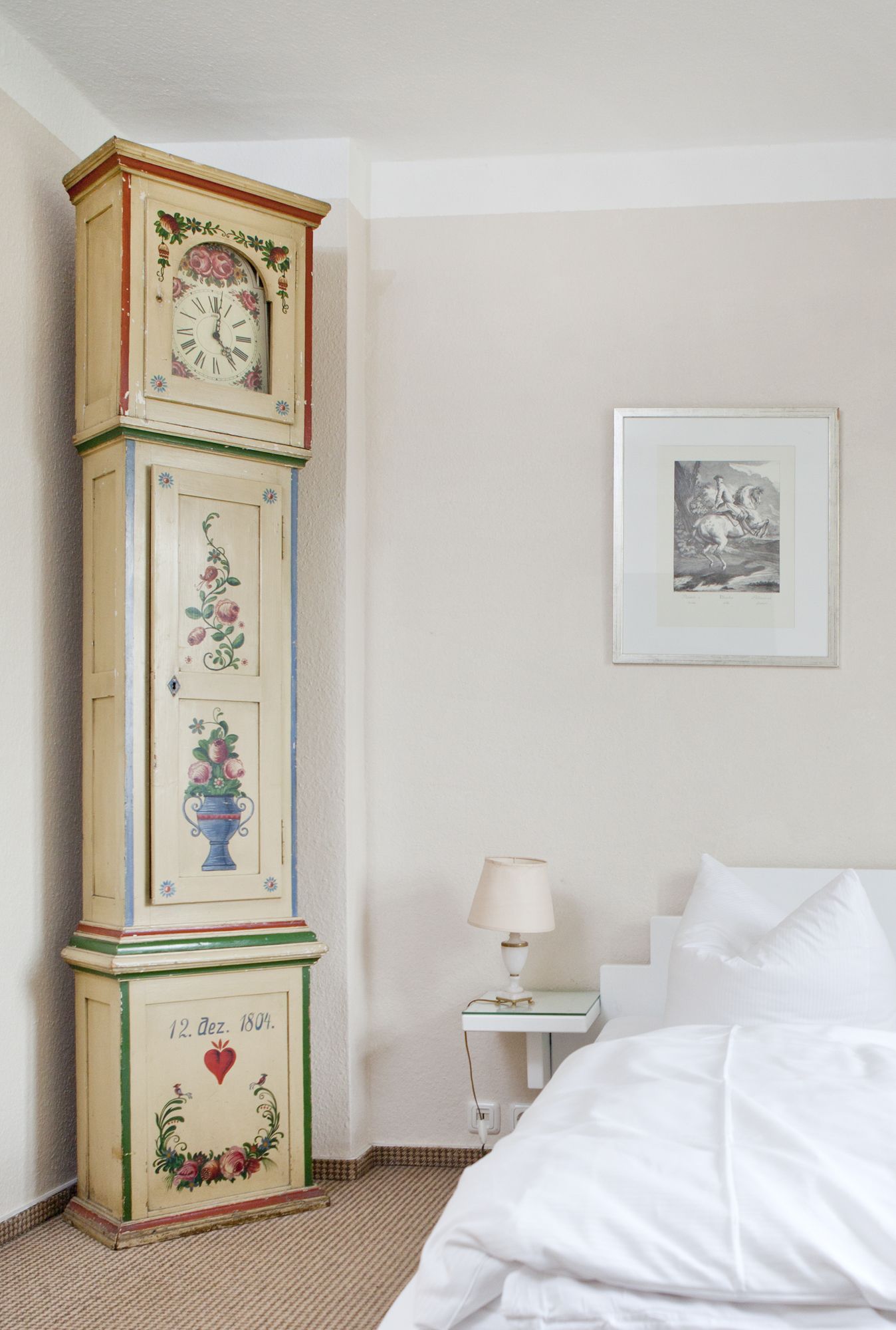 Un dormitorio con un reloj alto y ornamentado, un cuadro en la pared, una mesita de noche con una lámpara y una cama con ropa blanca.