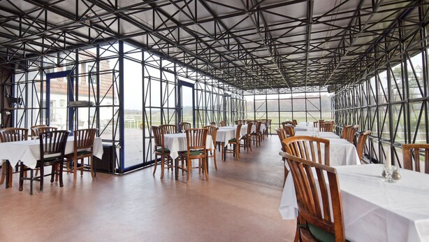 Un comedor interior con mesas y sillas, un techo alto con vigas metálicas y grandes ventanas que ofrecen vistas al exterior.