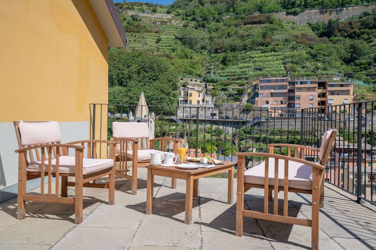 Un balcon avec des chaises en bois et une table préparée pour le petit-déjeuner, d’où l’on peut admirer des vignobles en terrasses et des bâtiments.