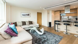 Een moderne woonkamer met een bank, een glazen salontafel en een vloerkleed. Er is een keukenhoek met barstoelen en een groot raam.