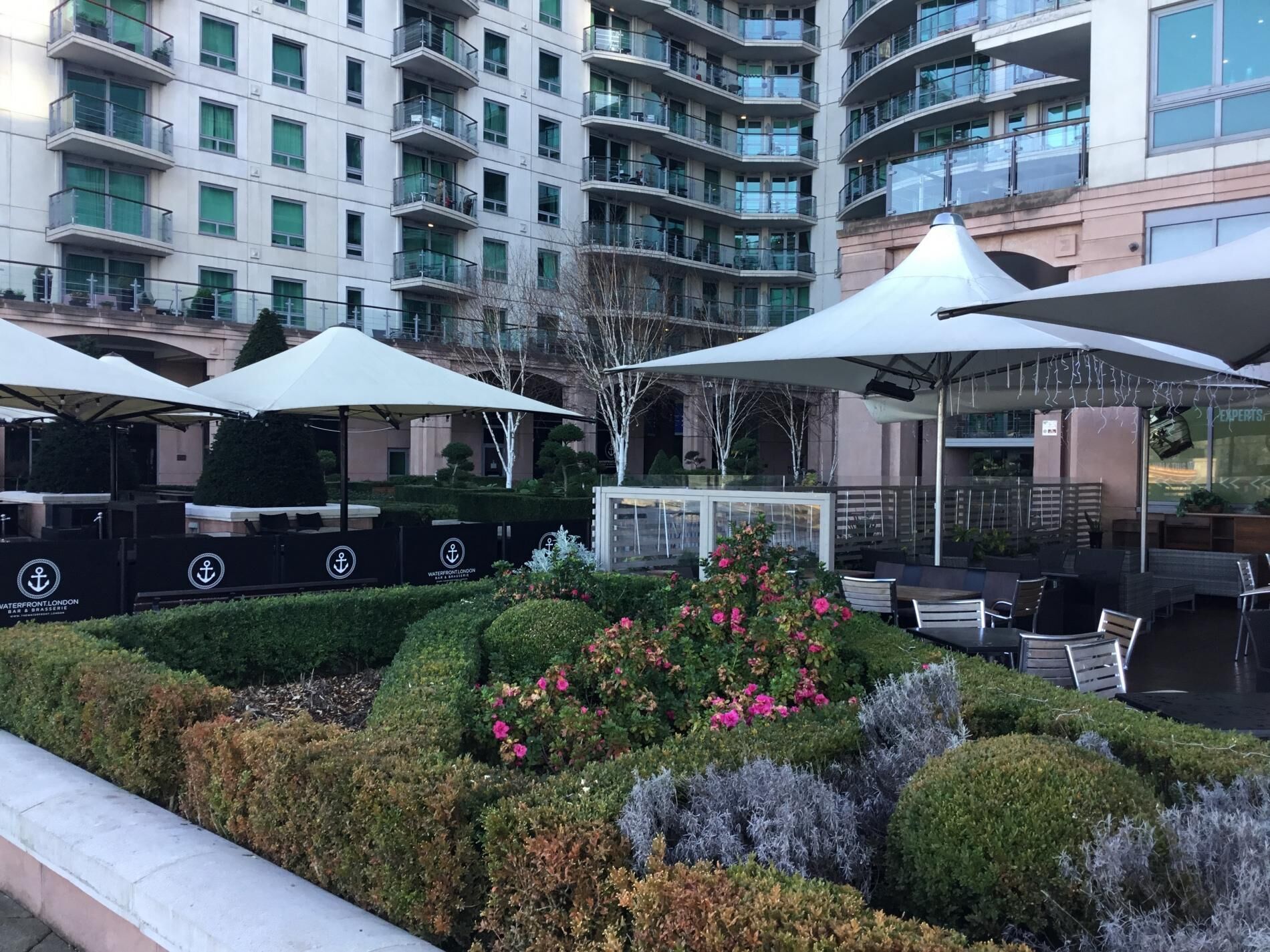 A modern outdoor seating area with umbrellas, a well-maintained garden, and a multi-story building in the background.
