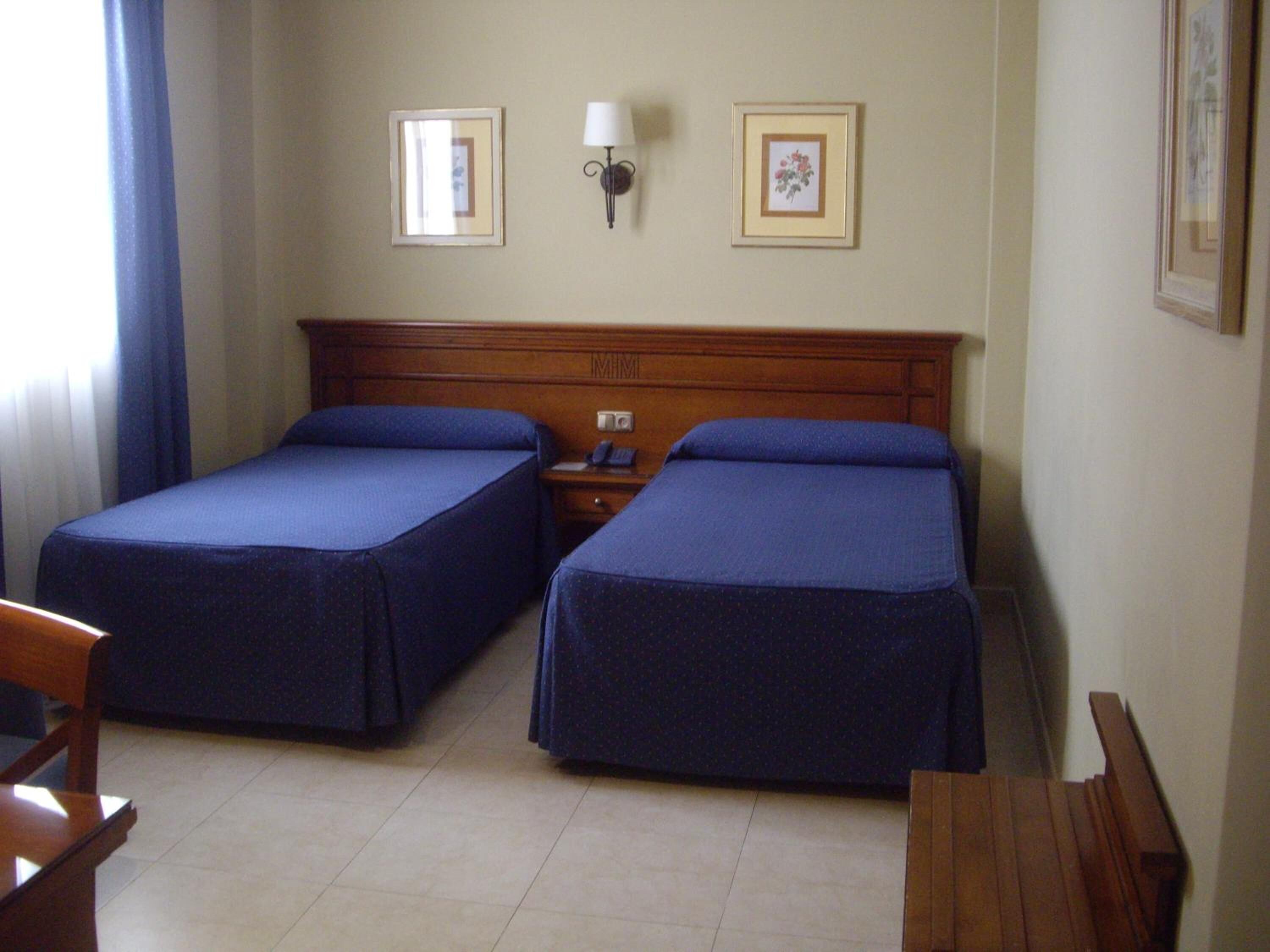 Una habitació d'hotel amb dos llits individuals, un panell de capçalera de fusta, una tauleta de nit amb una làmpada i dos quadres al mur.