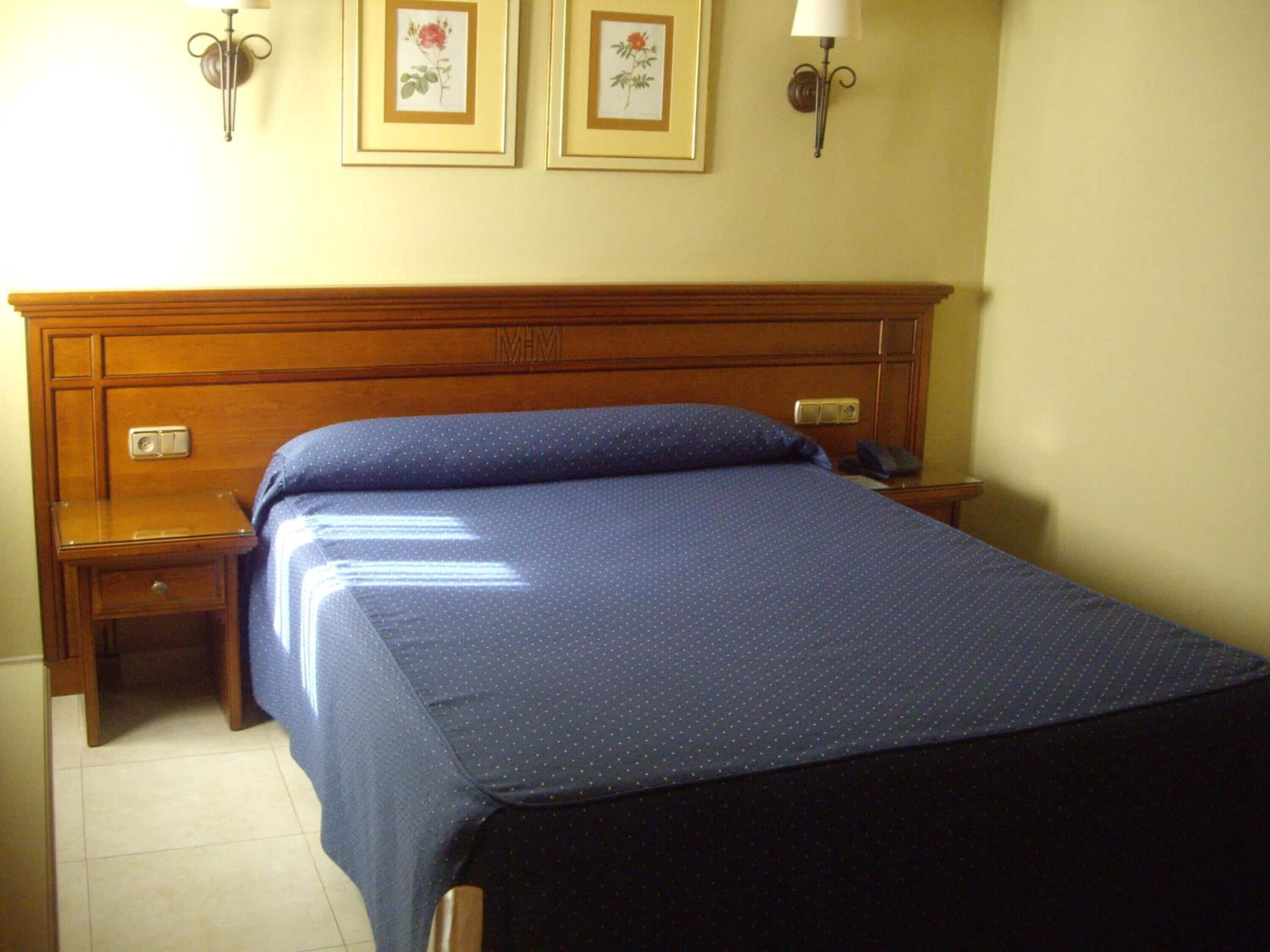 Una habitación de hotel estándar con cabecera de madera, una cama individual con colcha azul, dos mesitas de noche con lámparas y dos cuadros florales enmarcados en la pared.