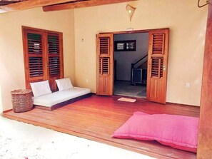 A room with a wooden floor, a bed with white bedding, a pink cushion, and wooden shutters on the windows.