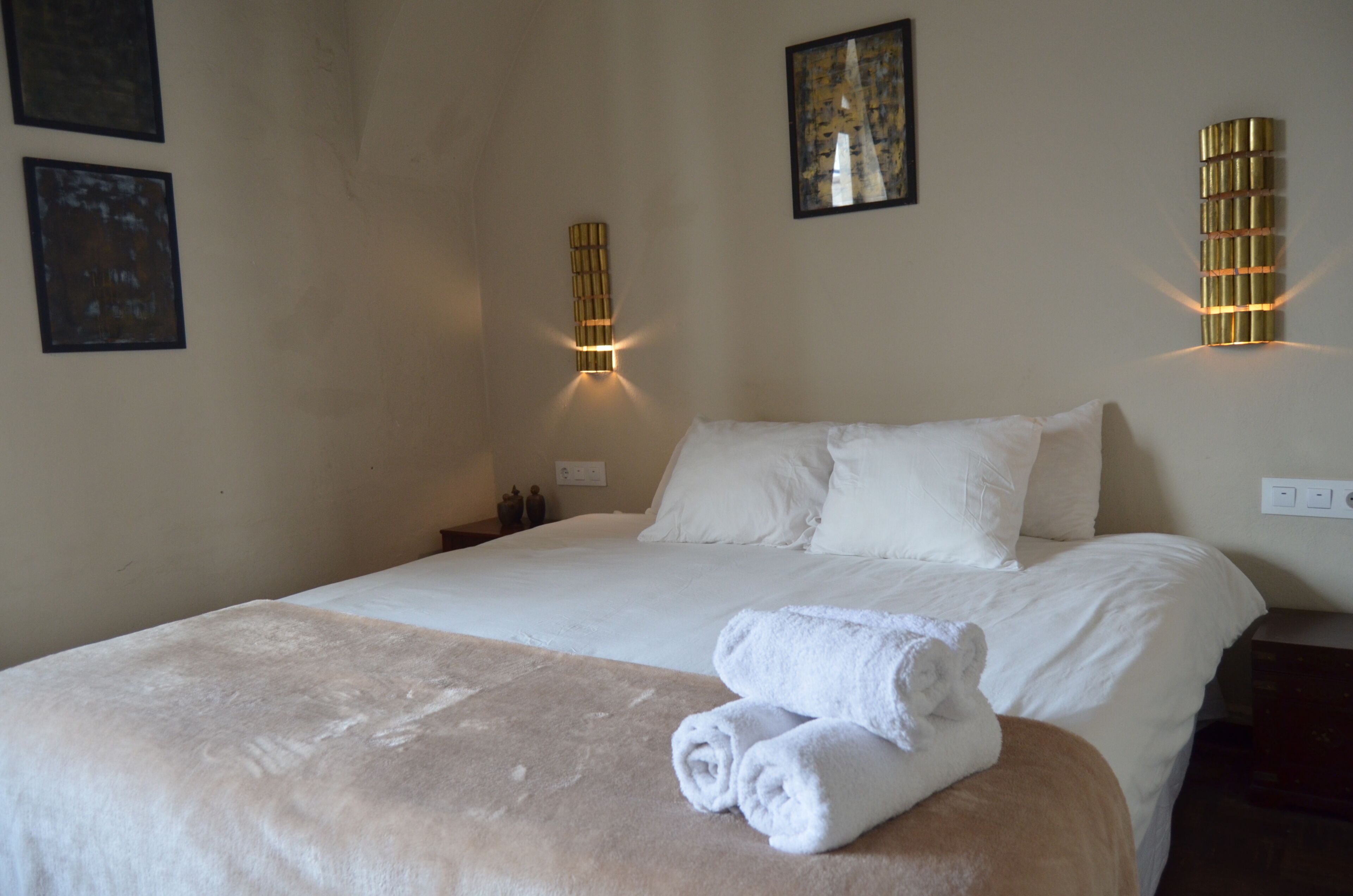 A neatly made bed with white linens and a beige blanket, two wall-mounted light fixtures, and two framed artworks on the wall.