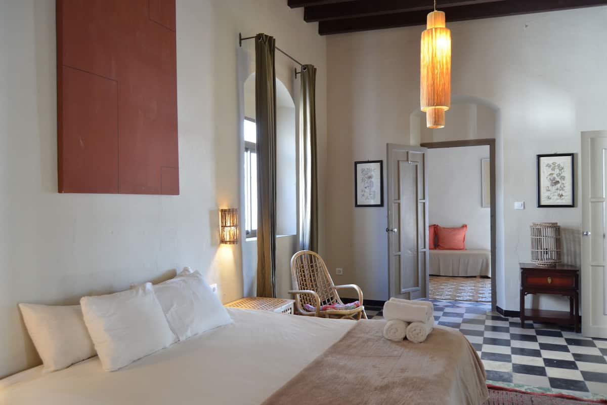 A hotel room with a large bed, a chair, a small table, and a hanging light fixture.