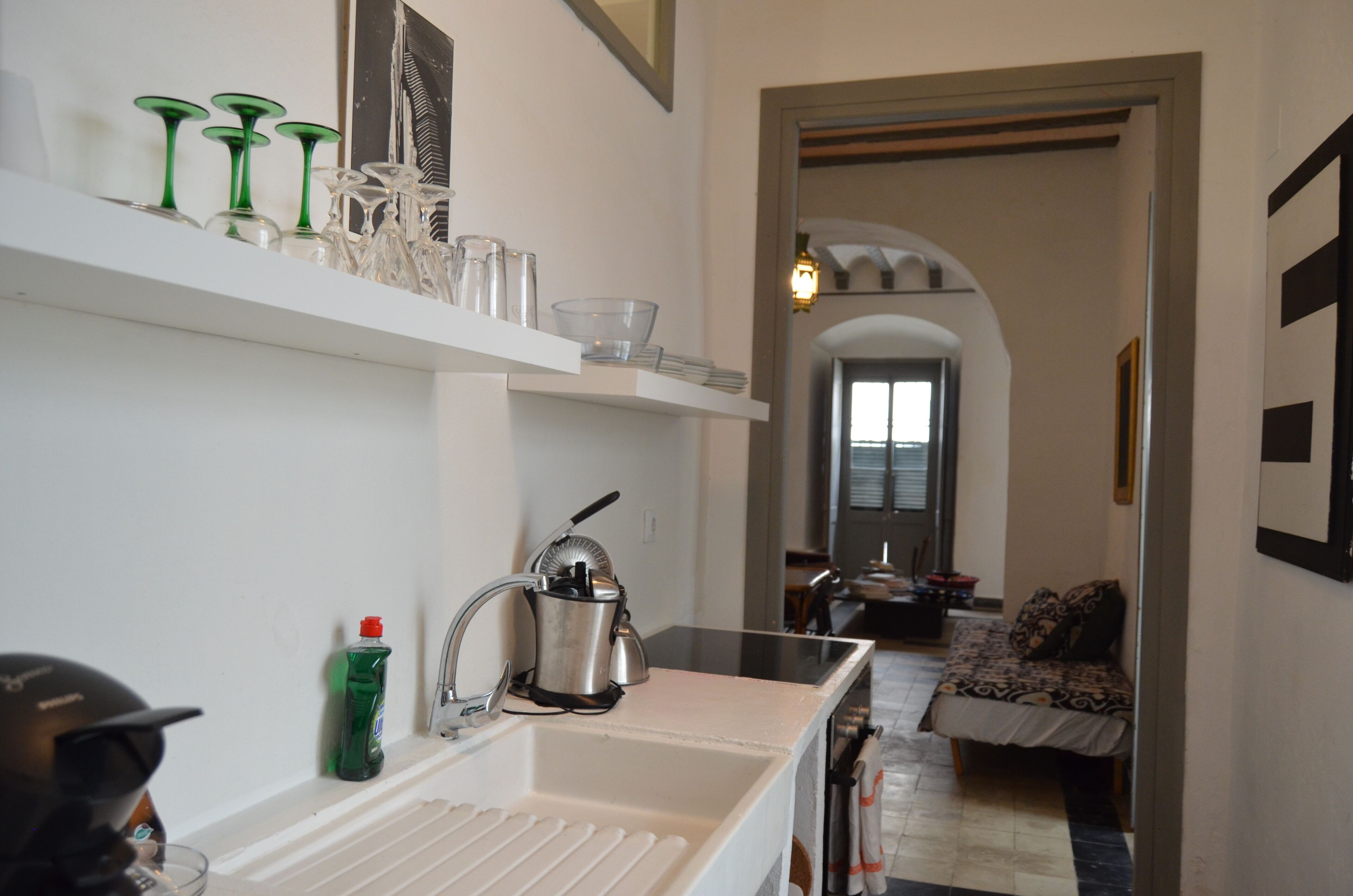 Una cocina con mostrador blanco, un lavamanos y una tetera. Hay una repisa con cristalería y una botella de detergente. Un pasillo conduce a un comedor con mesa y sillas.