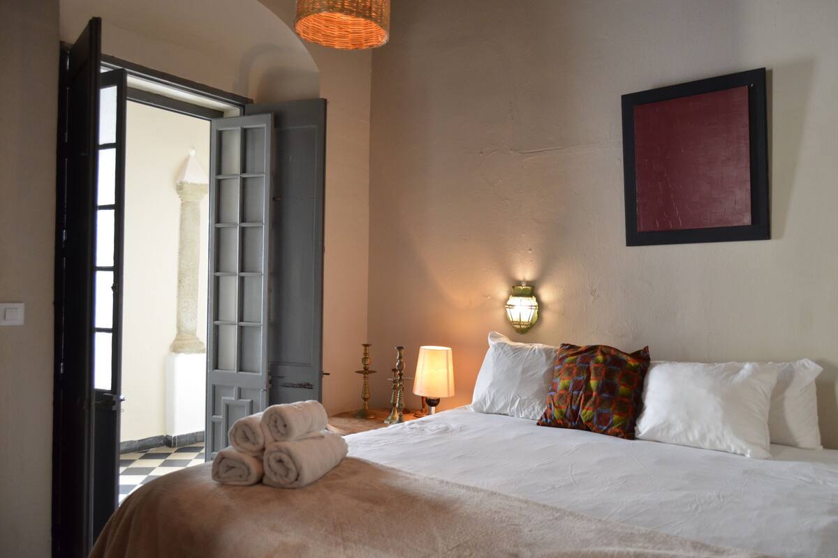 A hotel room with a bed, a nightstand with a lamp, a wall-mounted light, and a door leading to another room.