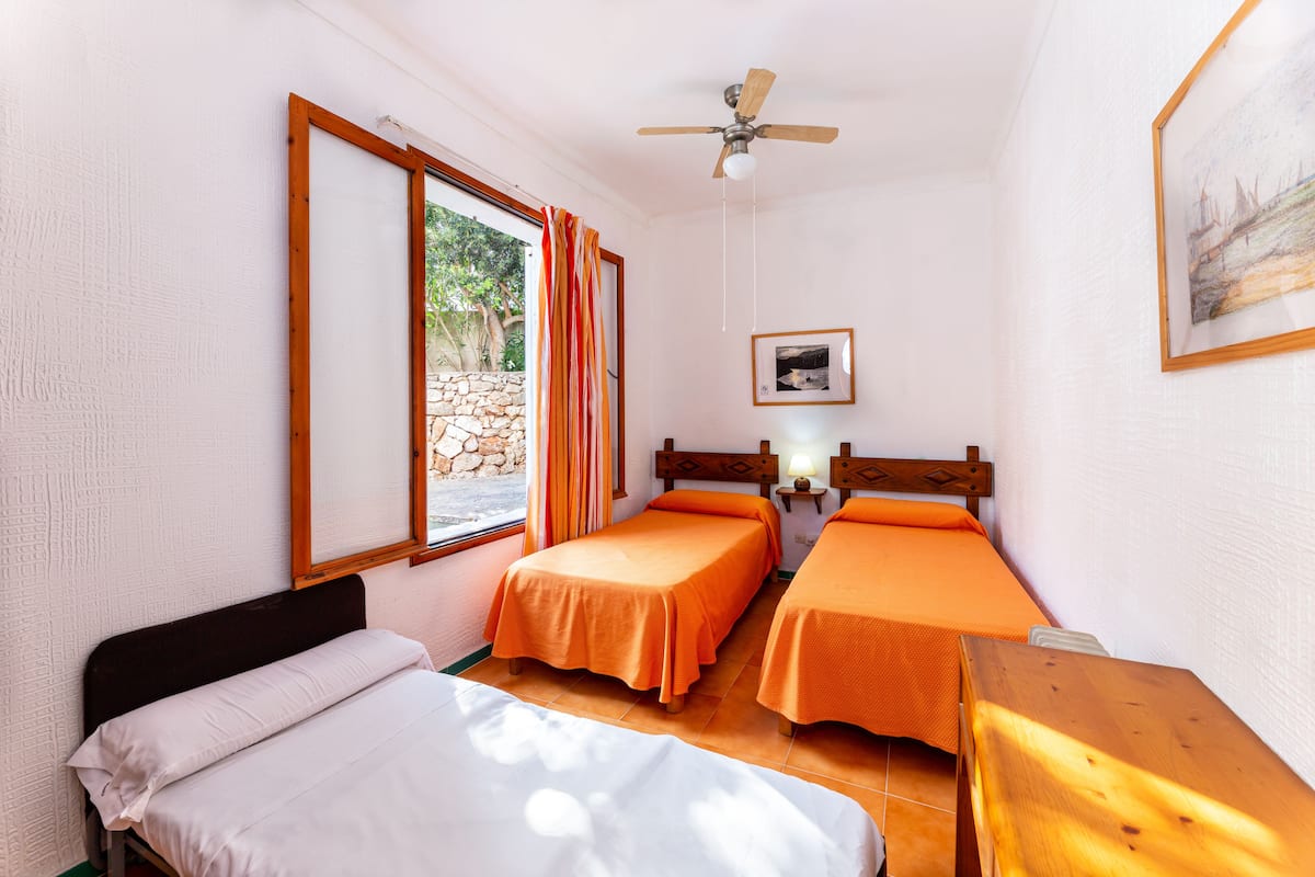 Una habitación con dos camas individuales, una cama más grande, una mesa de madera, un ventilador de techo, una ventana con cortinas y un cuadro en la pared.