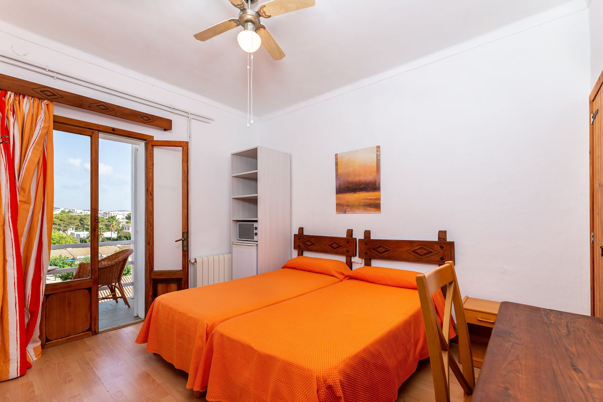 Una habitación con una cama, una mesa de madera, una silla, un ventilador de techo y vistas a través de una puerta de cristal.