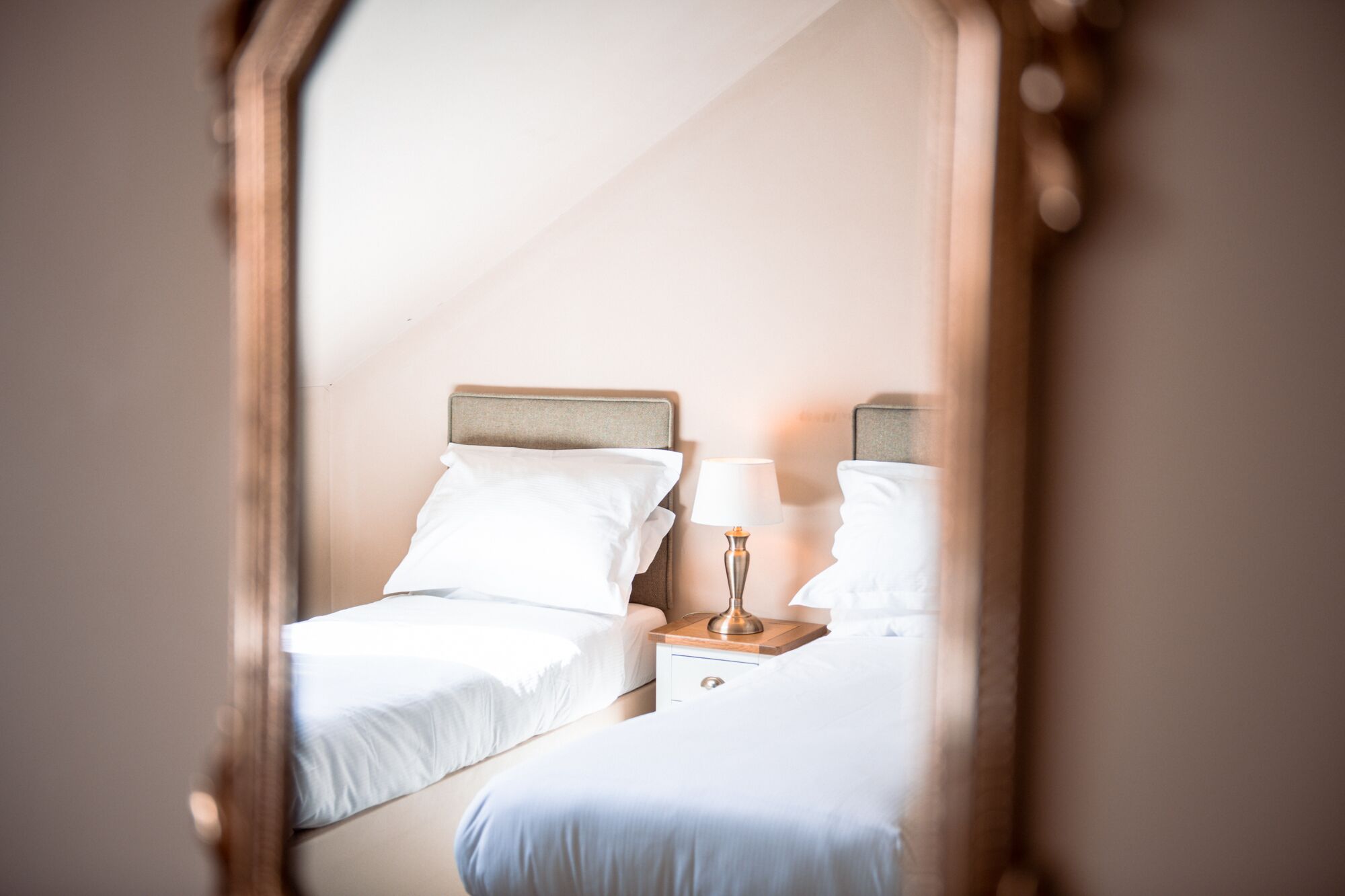 A bedroom with two beds, a nightstand with a lamp, and a mirror reflecting the room.