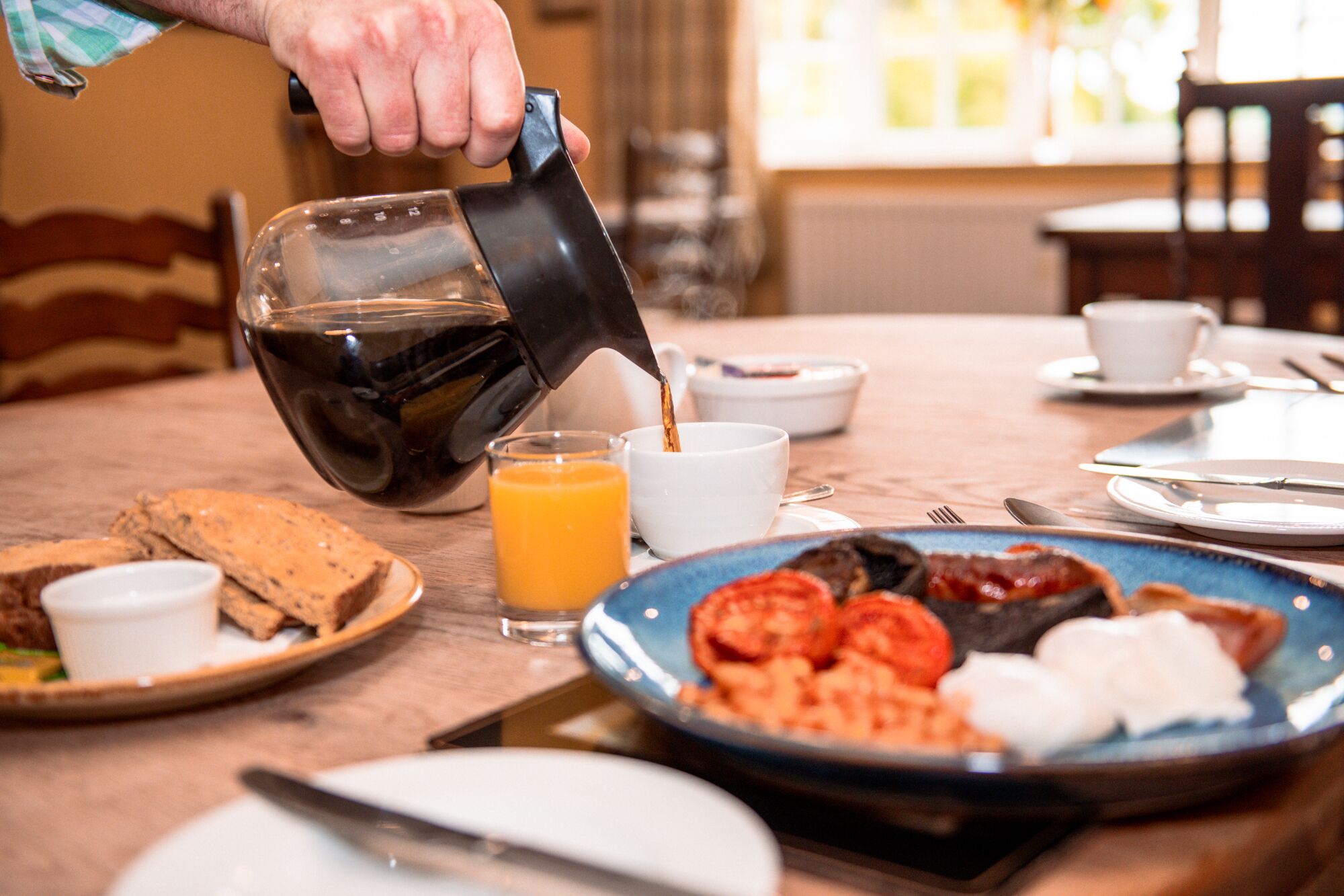 Un desayuno con una cafetera sirviendo café en una taza, un plato con huevos, tomates y una rebanada de pan.