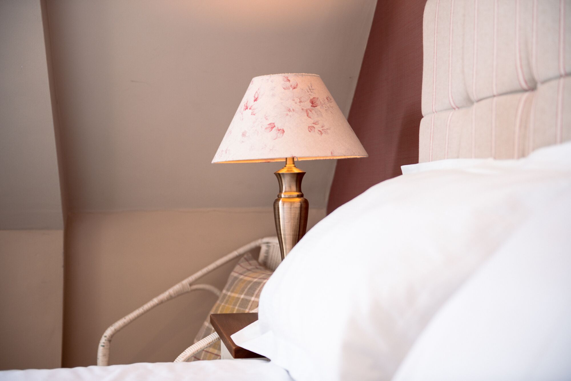 A bed with white bedding, a lamp with a floral lampshade, and a wicker chair with a plaid cushion.