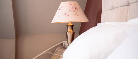 A bed with white bedding, a lamp with a floral lampshade, and a wicker chair with a plaid cushion.