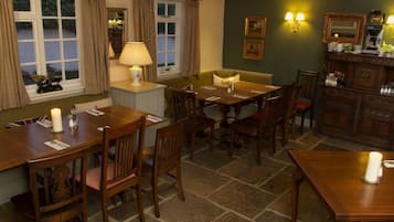 A dining area with wooden tables and chairs, a lamp, and a cabinet with a mirror and framed pictures.