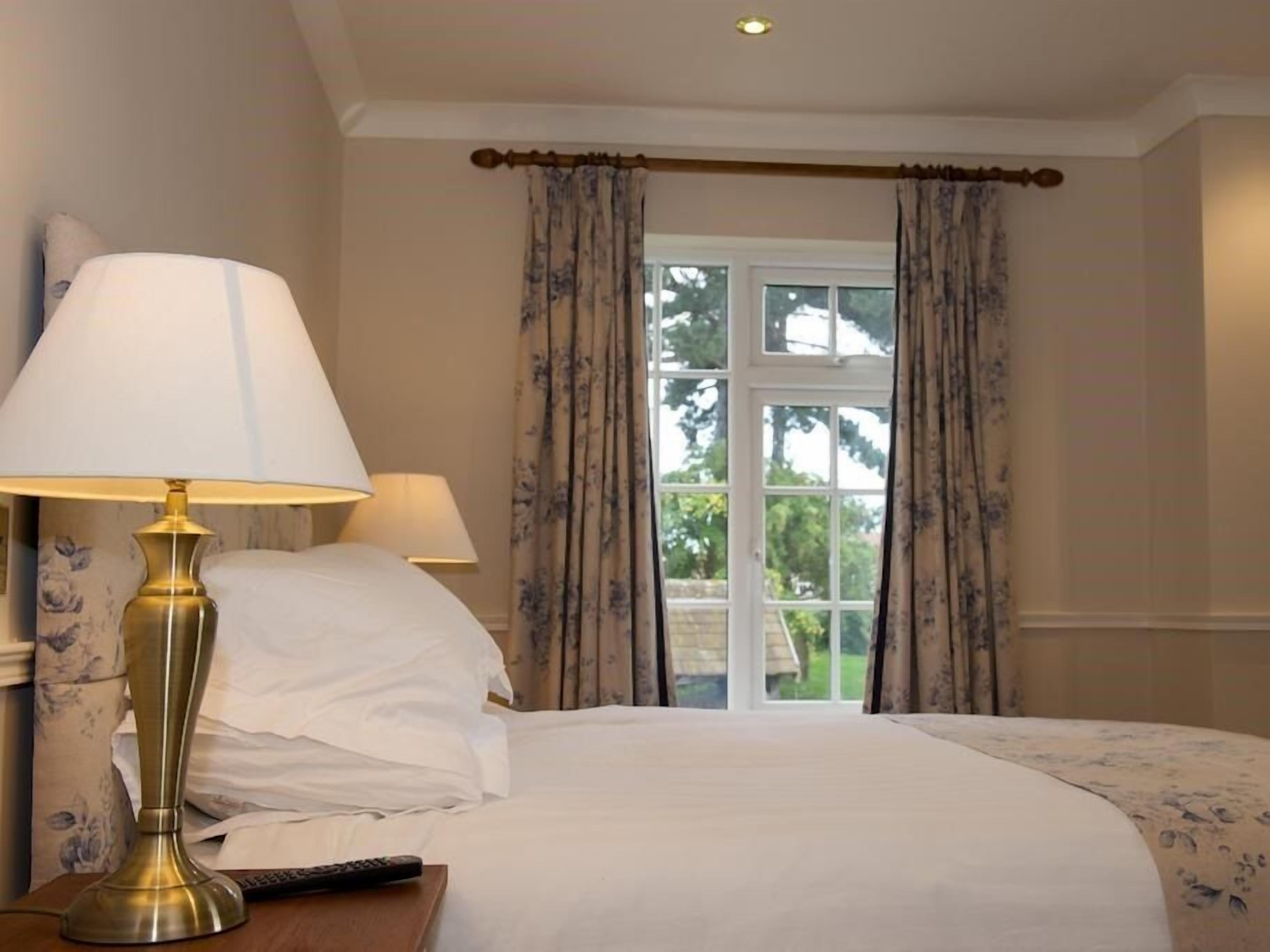 A hotel room with a bed, two bedside lamps, a remote control, and a window with curtains.