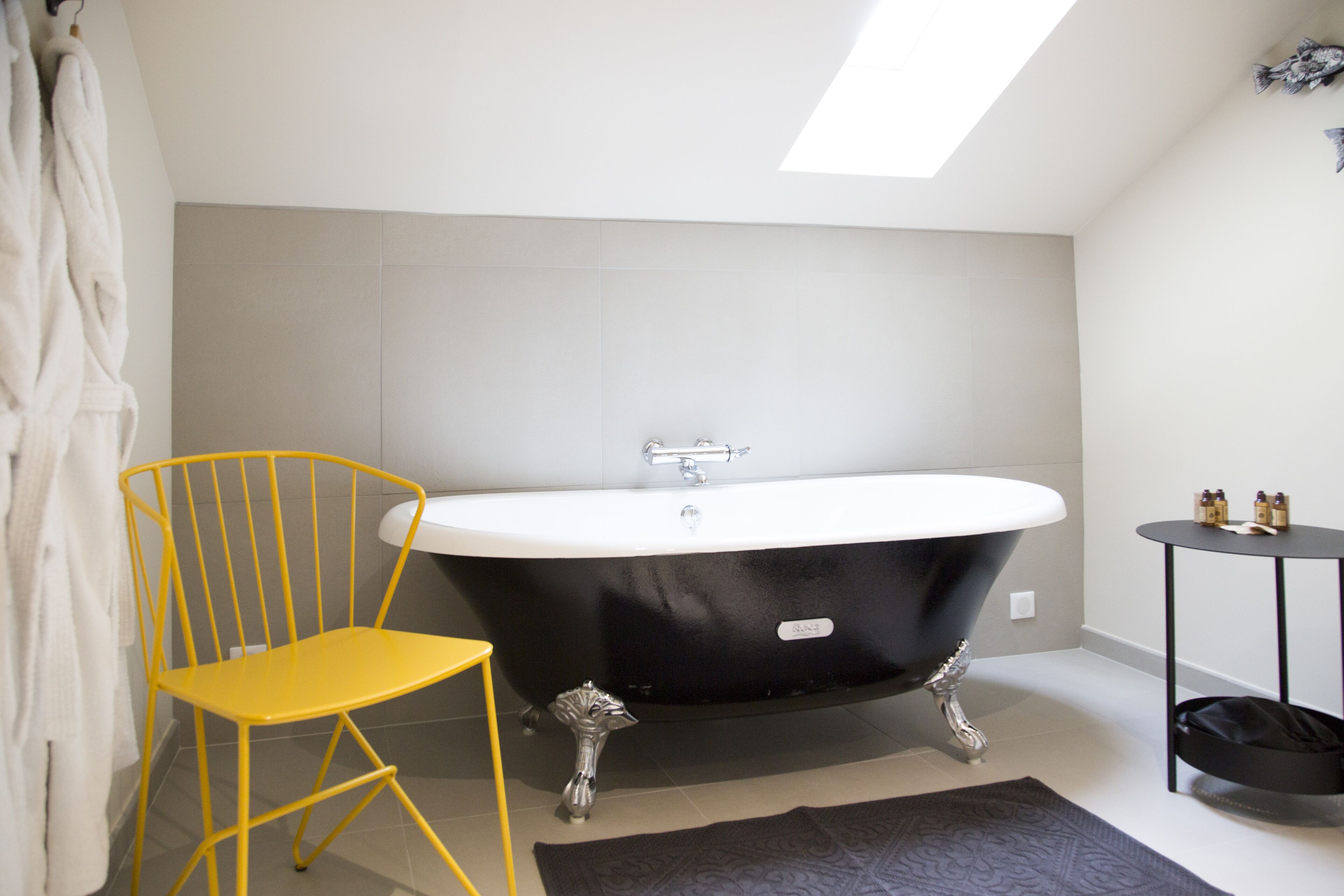 A modern bathroom with a freestanding bathtub, a yellow chair, and a small round table with toiletries.