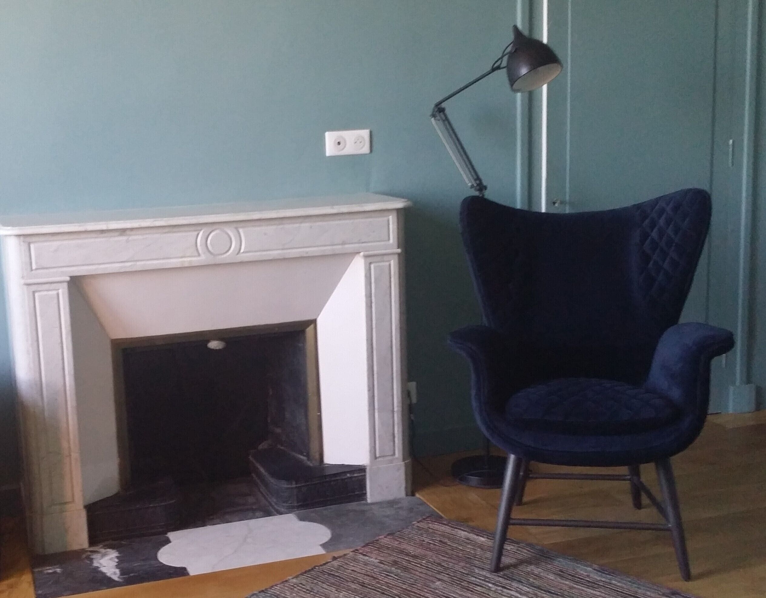 Una habitación con chimenea blanca, un sillón azul oscuro y una lámpara de pie.