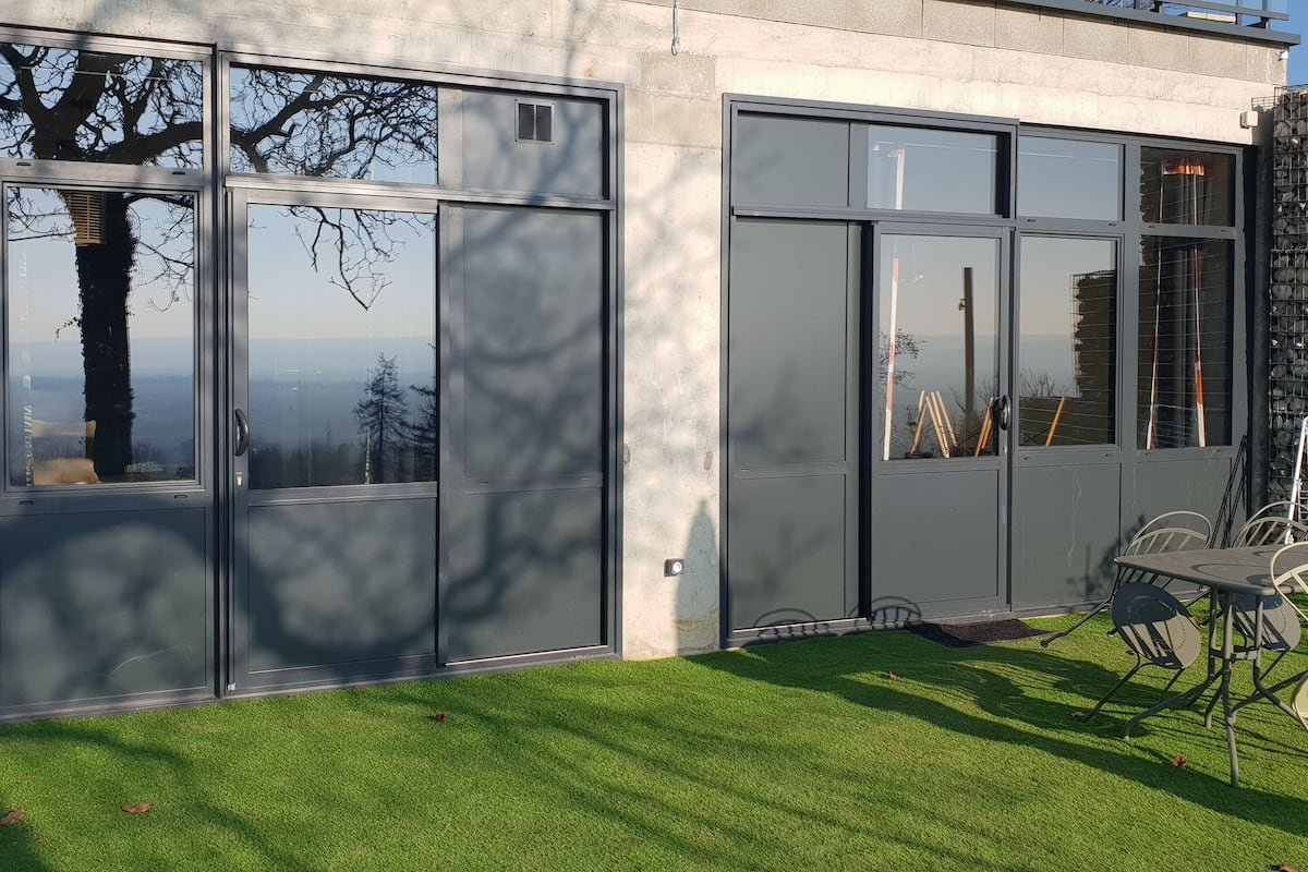 A modern building with large glass windows, a stone wall, and a grassy lawn with outdoor furniture.
