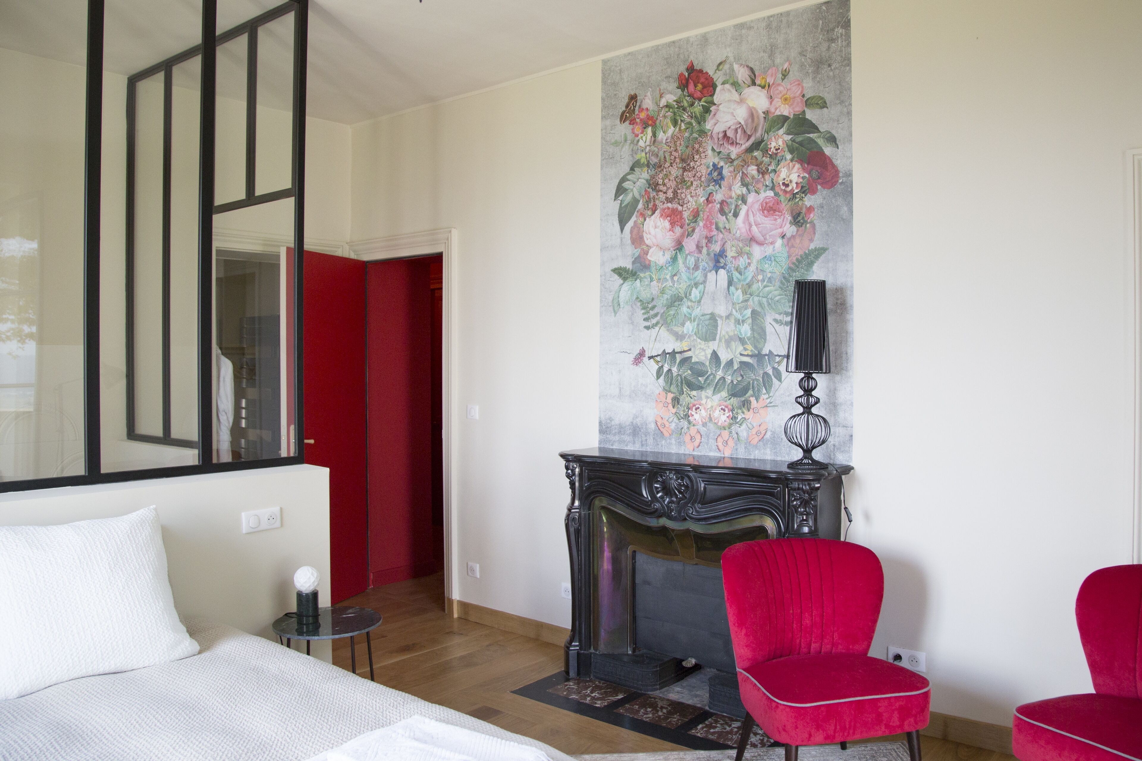A bedroom with a bed, a red chair, a fireplace, and a large floral painting on the wall.