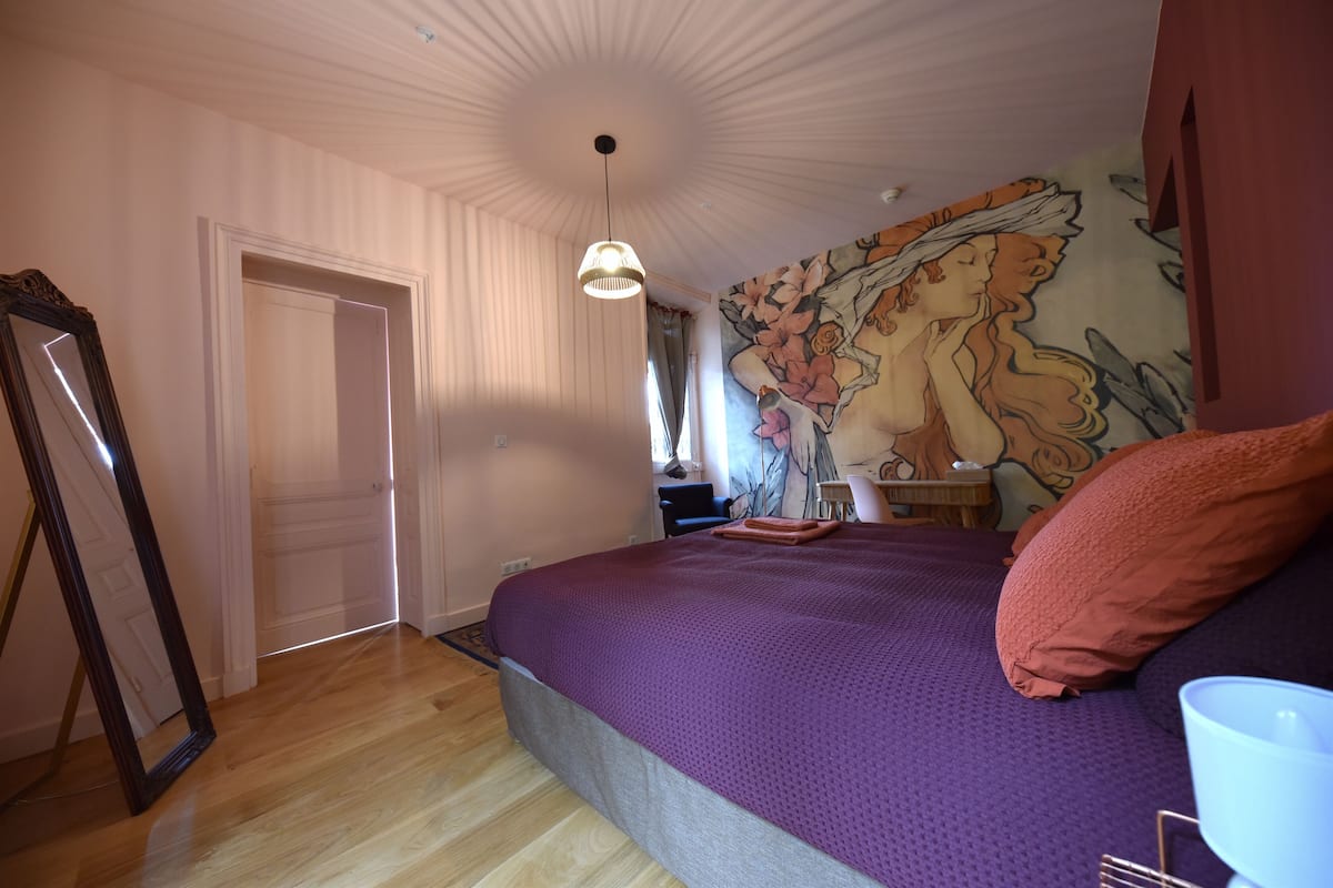 A bedroom with a large bed, a wooden floor, a ceiling with a unique design, and a mural on the wall.