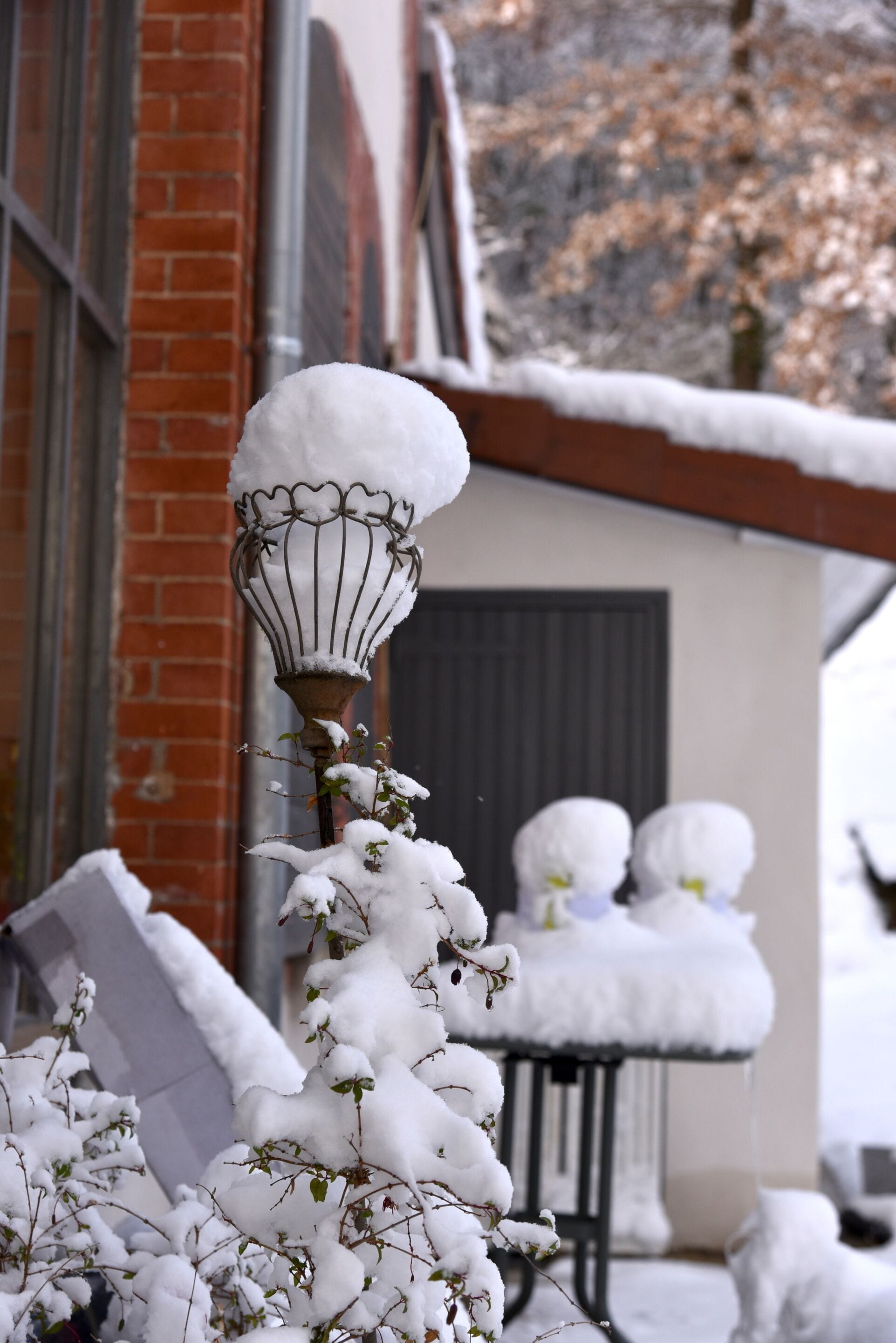 A snow-covered street lamp and plants in front of a building with a brick facade and a window.
