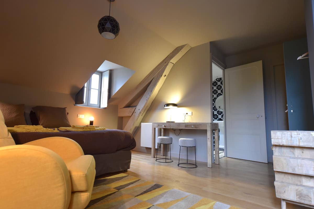 A cozy loft bedroom with a sloped ceiling, a bed, a desk with two stools, and a staircase.