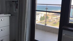 A room with a large window overlooking a coastal area, a kitchenette with a built-in oven, and a radiator. - Sea View Studio Mamaia (Constanta)