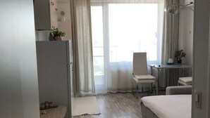 A hotel room with a bed, a desk, a chair, a small table, a refrigerator, and a window with curtains. - Sea View Studio Mamaia (Constanta)