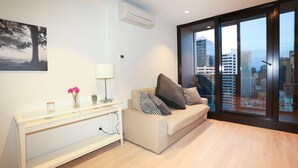 A modern living room with a sofa, a side table, a lamp, and a city view through large windows. - Central But Quiet Apartment (Melbourne)