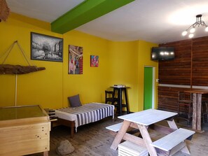 A room with a yellow wall, a bed with a striped bedspread, a small table with books, a wooden bench, and a television mounted on the wall.