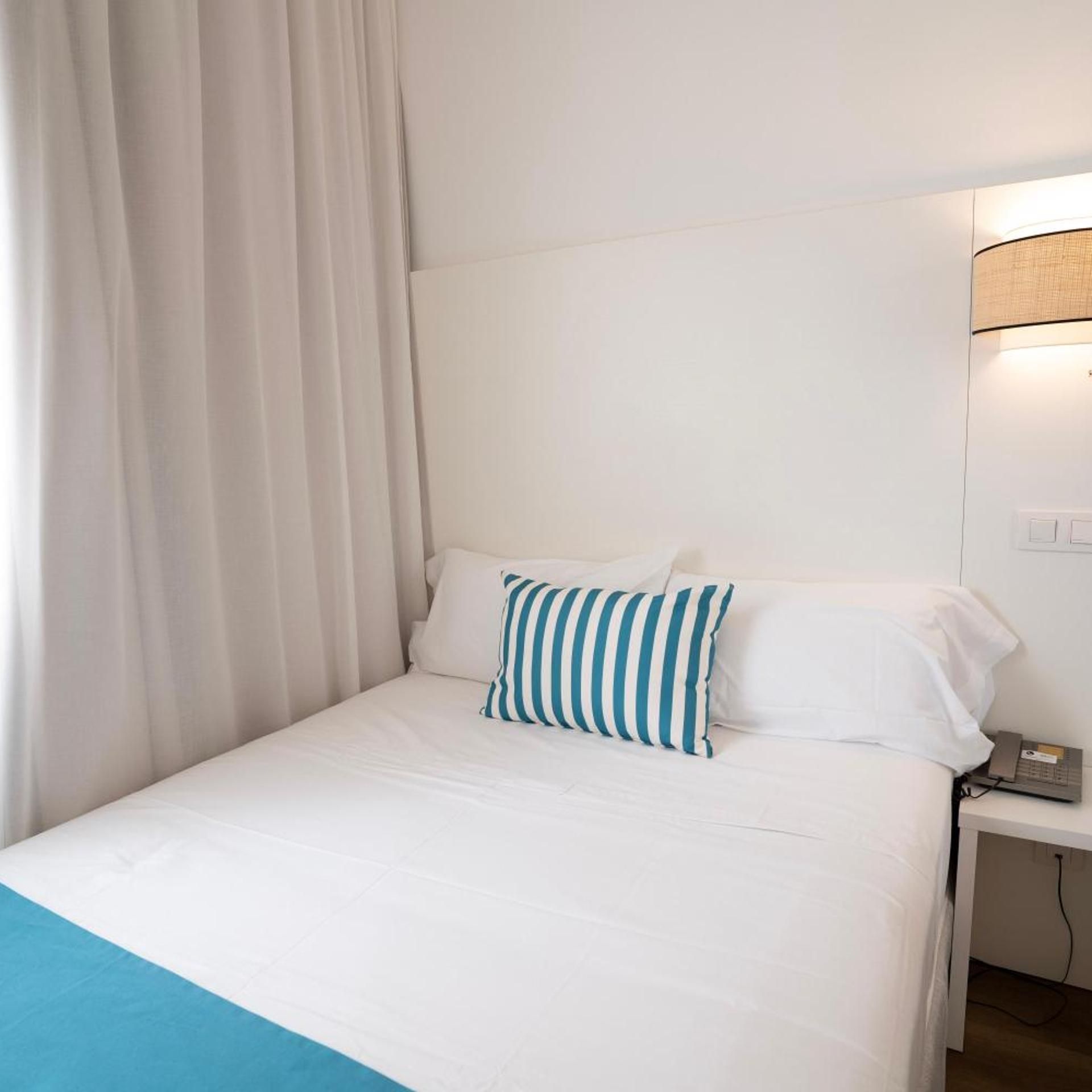 Quarto de hotel com cama, mesa de cabeceira, luminária e cortina.