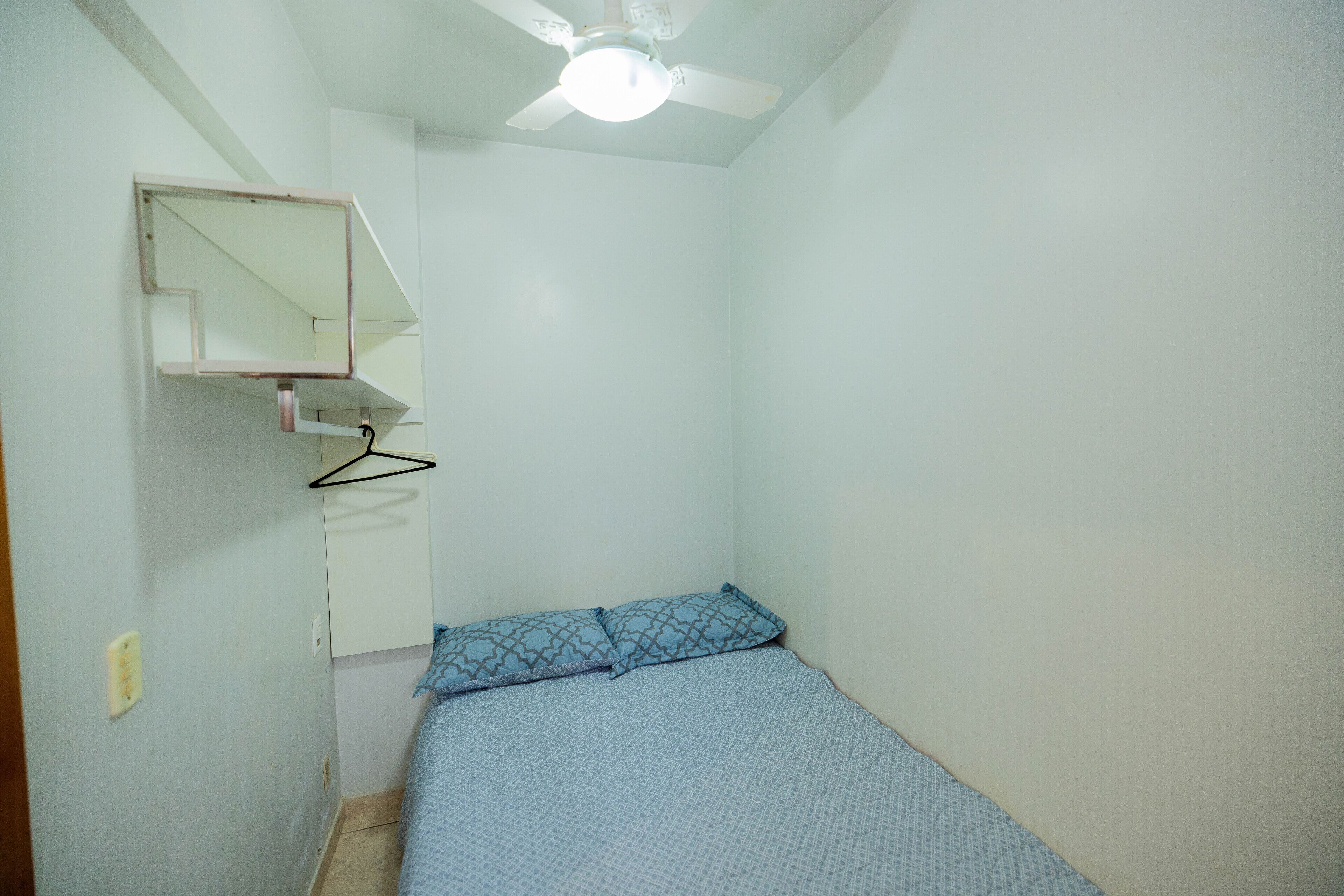 Une petite pièce, peu meublée, avec un lit simple, une étagère fixée au mur et un ventilateur de plafond.